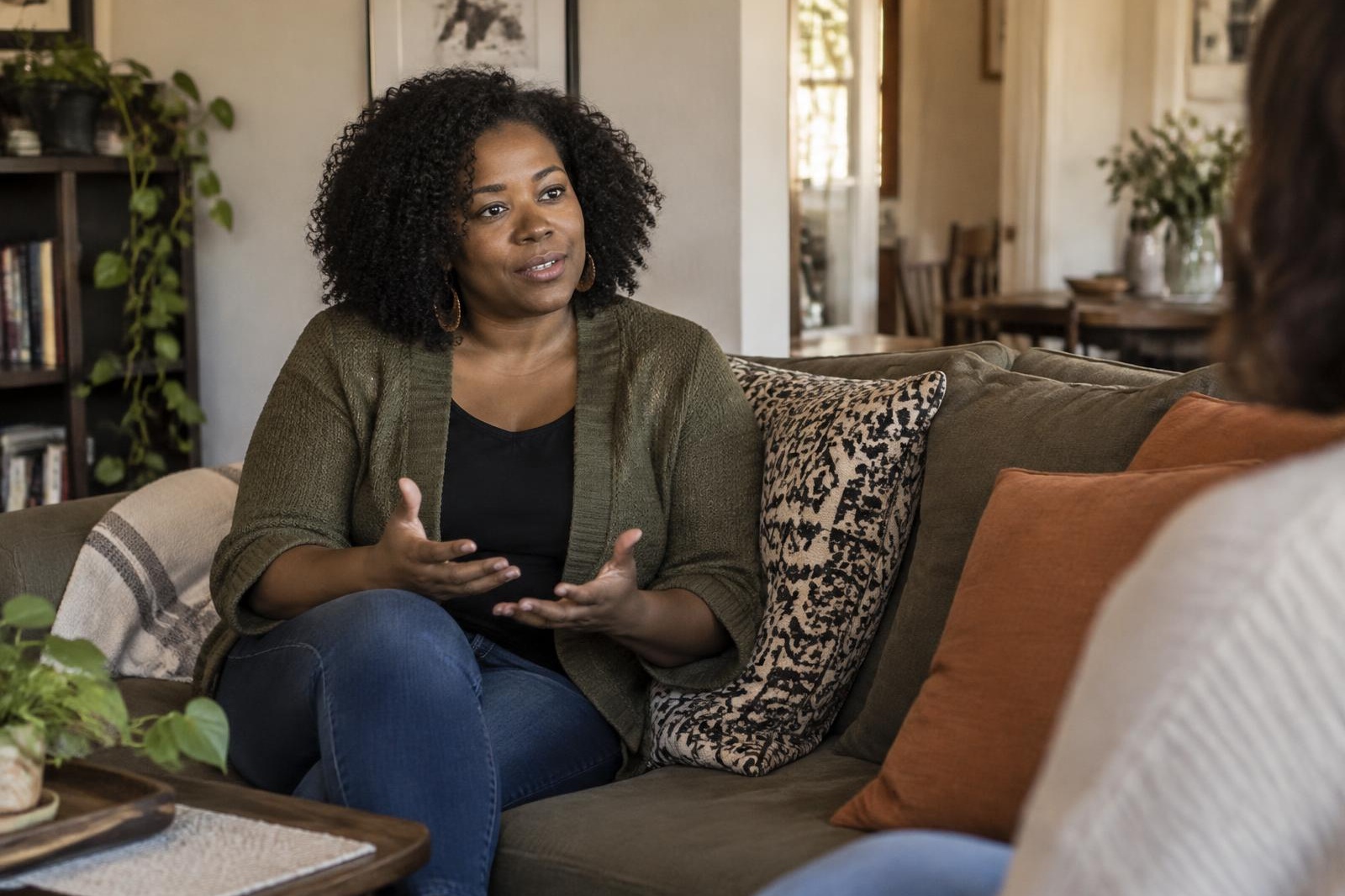 A woman with natural curly hair talking thoughtfully while seated on a couch in a warm living room