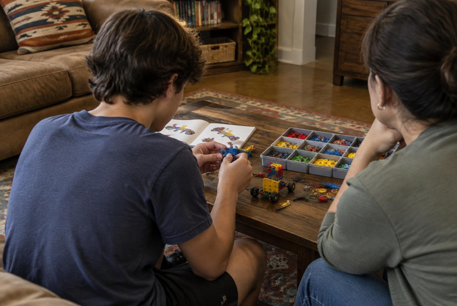 A teen building a small model while an adult listens nearby